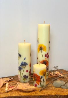 three candles with flowers painted on them sitting on a wooden table next to dried leaves