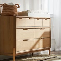 a wooden dresser sitting in front of a window next to a white bed with a basket on top of it