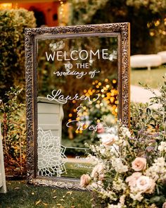 a welcome sign in front of a mirror with flowers and greenery around it on the grass