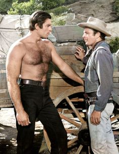 two shirtless men standing next to each other in front of an old wooden wagon