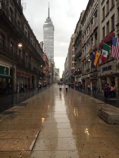 people are walking down an empty city street in the rain with buildings and flags on either side