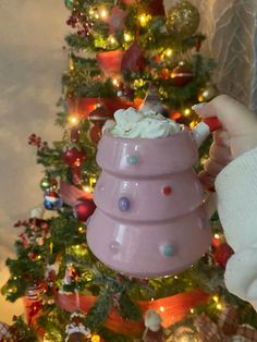 a hand holding a pink cup with white frosting on it in front of a christmas tree