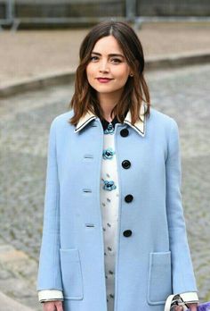 a woman in a blue coat and white shirt is holding a purple handbag while standing on the street