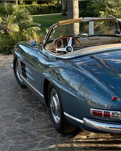 an old blue sports car parked in front of a palm tree on a stone driveway