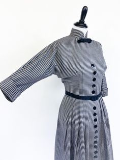 "A fabulously chic vintage day dress from the early 1950s! Medium weight cotton blend in horizontal black, white and gray stripes. Fitted waist and bloused bustline with cuffed ¾ length sleeves cut in one with the bodice. High Nehru collar accented with tailored grosgrain ribbon bow. A-line skirt with gathers at each hip, front and back. Dress closes up the front with a row of 22 buttons that extend from bust to hem. Small shoulder pads and short metal zipper at back neck. Unlined, missing origi Fitted Dress With Striped Collar For Daywear, Vintage Striped Dresses For Work, Vintage Striped Dress For Work, Vintage Striped Workwear Dresses, Vintage Long Sleeve Striped Dresses, Retro A-line Dress With Button Closure, Striped Retro Dresses With Buttons, Vintage Striped Dresses For Daywear, 1950s Style A-line Vintage Dress With Buttons