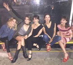 four women sitting on a bench in front of a bus at night time, with their arms up