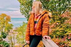 a woman in an orange jacket is standing on a wooden railing by the water and trees