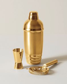 a gold cocktail shaker, cup and spoon on a white surface with a silver background