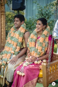 two people sitting on a swing with flowers in their hair and garlands around their necks