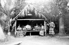 an old black and white photo of three women sitting in front of a small hut