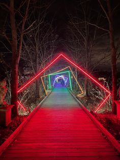 a wooden walkway with red and green lights on it at night, leading to a tunnel in the woods