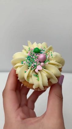 a hand holding a cupcake with white frosting and green sprinkles