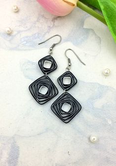 three square shaped earrings sitting on top of a table next to a pink tulip