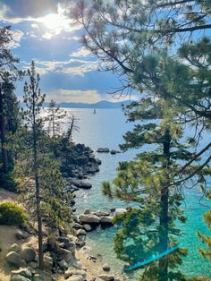 the sun shines through the clouds over some trees and rocks by the water's edge