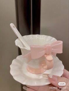 a hand holding a white cup and saucer with pink bow on the top, in front of a mirror