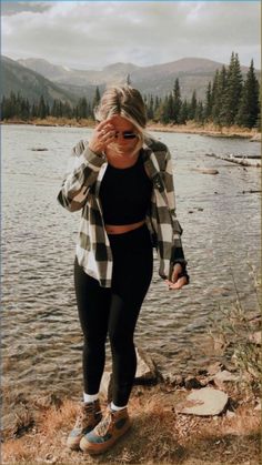 a woman standing next to a body of water wearing leggings and a checkered shirt