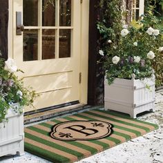 two planters with flowers are on the front porch next to a door mat that says o c