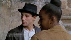 a man and woman standing next to each other near a stone wall with a black top hat on