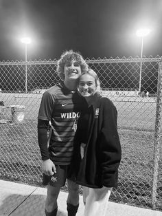 a man and woman standing next to each other in front of a fence at night