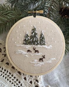 a cross stitch christmas ornament hanging on a tree branch with pine trees and snow