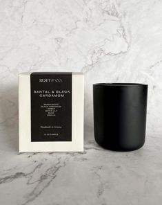 a black candle sitting next to a white box on a marble counter top, with the packaging in front of it