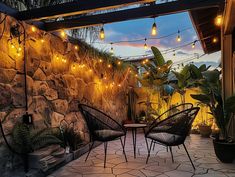 an outdoor patio with string lights strung from the ceiling and chairs on the ground, next to a stone wall