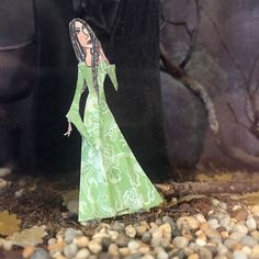 a paper doll standing in front of a tree with rocks and gravel on the ground