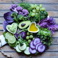 an assortment of vegetables are arranged on a wooden surface with dressing in the center and olives, broccoli, cucumbers, grapes, avocados, and more