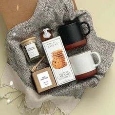 two coffee mugs, honey and cookies are on a blanket next to a box