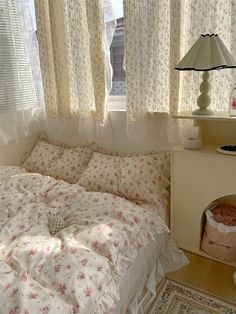 a white bed sitting under a window next to a night stand and table with a lamp on it