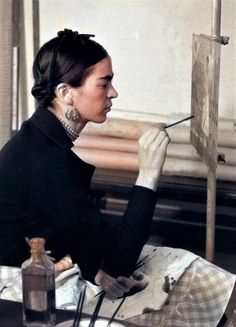 a woman sitting in front of a easel holding a paintbrush and looking at something