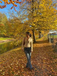 a woman walking down a leaf covered path next to a tree with yellow leaves on it