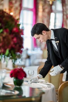 a man in a tuxedo and white gloves is setting a table with wine glasses