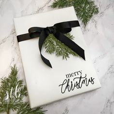 a white box with a black ribbon and merry christmas lettering on it sitting next to evergreen branches