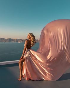 a woman in a pink dress sitting on a ledge next to the ocean and looking off into the distance