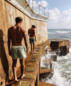 two men are walking along the edge of a pier by the ocean with waves crashing in front of them