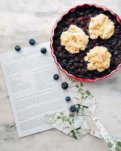 blueberry cobbler in a red bowl next to an open recipe book on a marble table