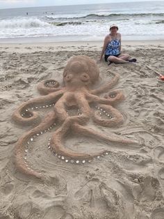 an octopus made out of sand on the beach with a woman sitting next to it