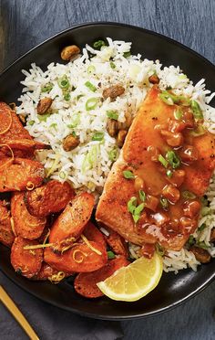 a black plate topped with rice and meat covered in sauce next to lemon wedges