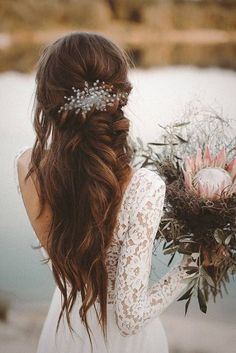 a woman with long hair holding a bouquet and wearing a white lace dress, looking at the water