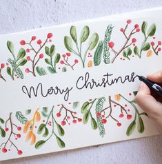 a person is writing merry christmas on a card with watercolor leaves and red berries