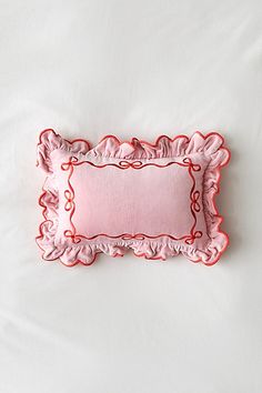 a pink pillow with ruffled edges on a white bed sheet that has red trim around the edge