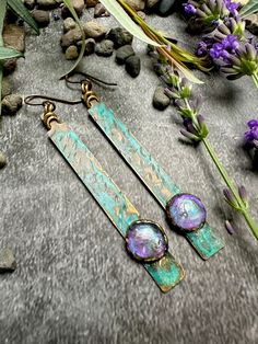 two earrings are laying on the ground next to some rocks and purple flowers with green leaves