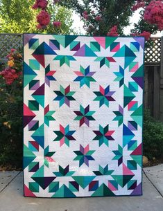 a colorful quilt is hanging on a fence in front of some trees and bushes with pink flowers