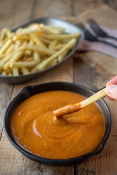 a person dipping french fries into a sauce in a black bowl on a wooden table