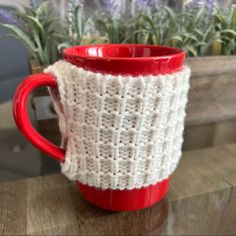 a red and white knitted coffee cup sitting on top of a wooden table next to plants