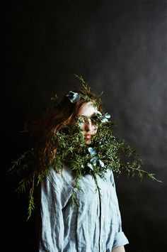 a woman with flowers on her head and branches around her neck, standing against a dark background