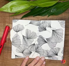 a person is drawing on a piece of paper next to some leaves and a red pen