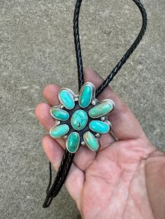 a hand holding a black leather cord with turquoise stones on it and a flower brooch