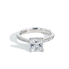 a princess cut diamond engagement ring with pave set diamonds on the band and an inscription that reads accordi